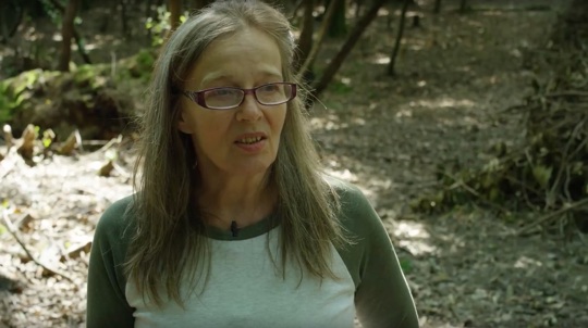 Woman with long hair and glasses in woodland talking to camera