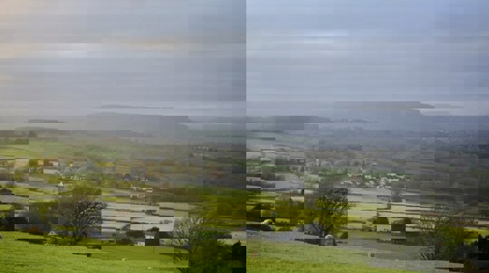 Green fields dotted with trees, hedges and white buildings under a dark misty sky