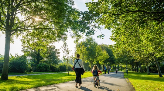 Woman walking with a child on a bicycle through a sunny park with lots of trees