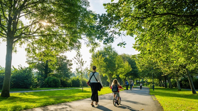 Woman walking with a child on a bicycle through a sunny park with lots of trees