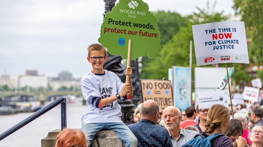 Smiling child with Woodland Trust placard and Tshirt sits on a wall as other campaigners walk by