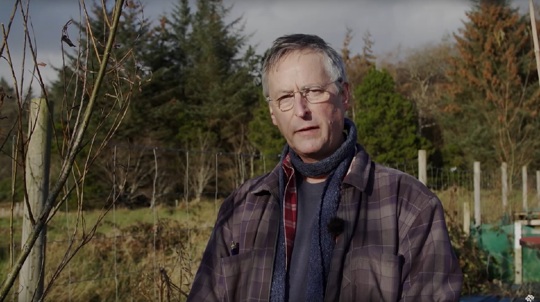 A man in glasses and a checked shirt stands among young trees talking to camera