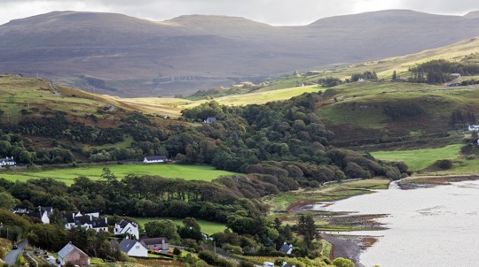 A wood sits over a hilly landscape close to the shoreline with white cottages in the foreground and mountains in the distance