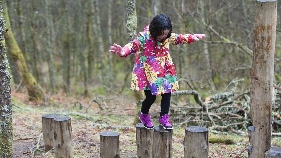 A young girl in a bright flowery coat walking across a row of tree stumps with arms outstretched