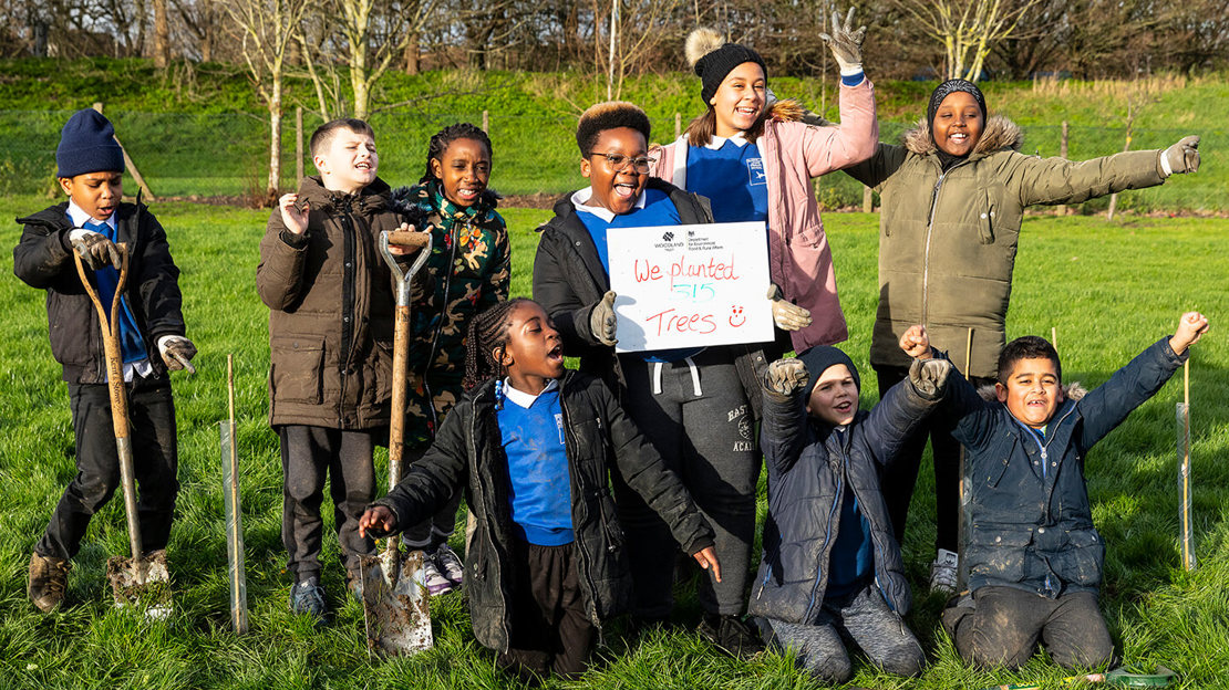 Group of schoolchildren smiling outdoors with spades and a sign that reads 'We planted 315 trees'