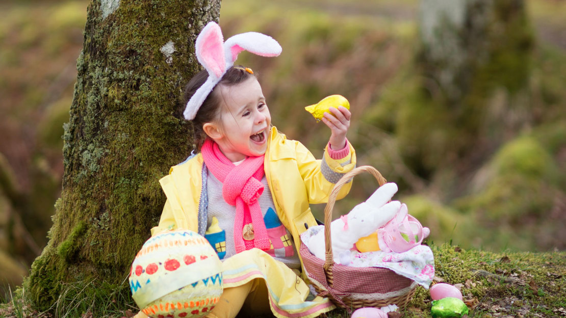 Smiling young girl wearing bunny ears, sitting on the woodland floor with a basket of Easter eggs