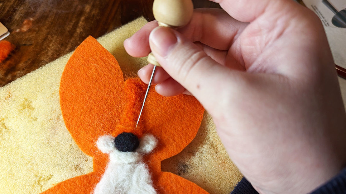 A hand holding a needle while felting wool to form a fox's face