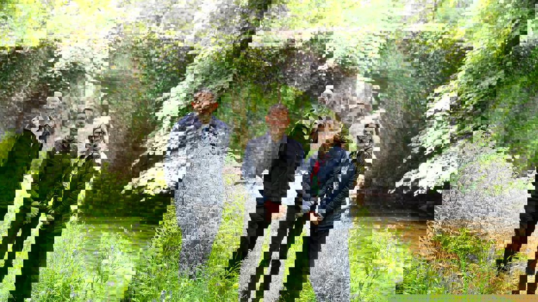 Three people stand in front of a stone bridge in leafy woodland on a sunny day