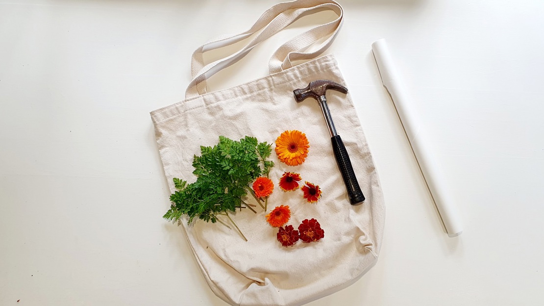 A canvas tote bag laid flat with flowers, leaves, baking paper and a hammer on top.