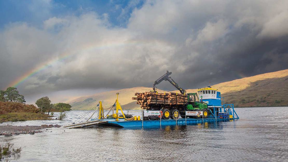 A blue barge carrying a green wagon loaded with logs floating on a loch under a rainbow