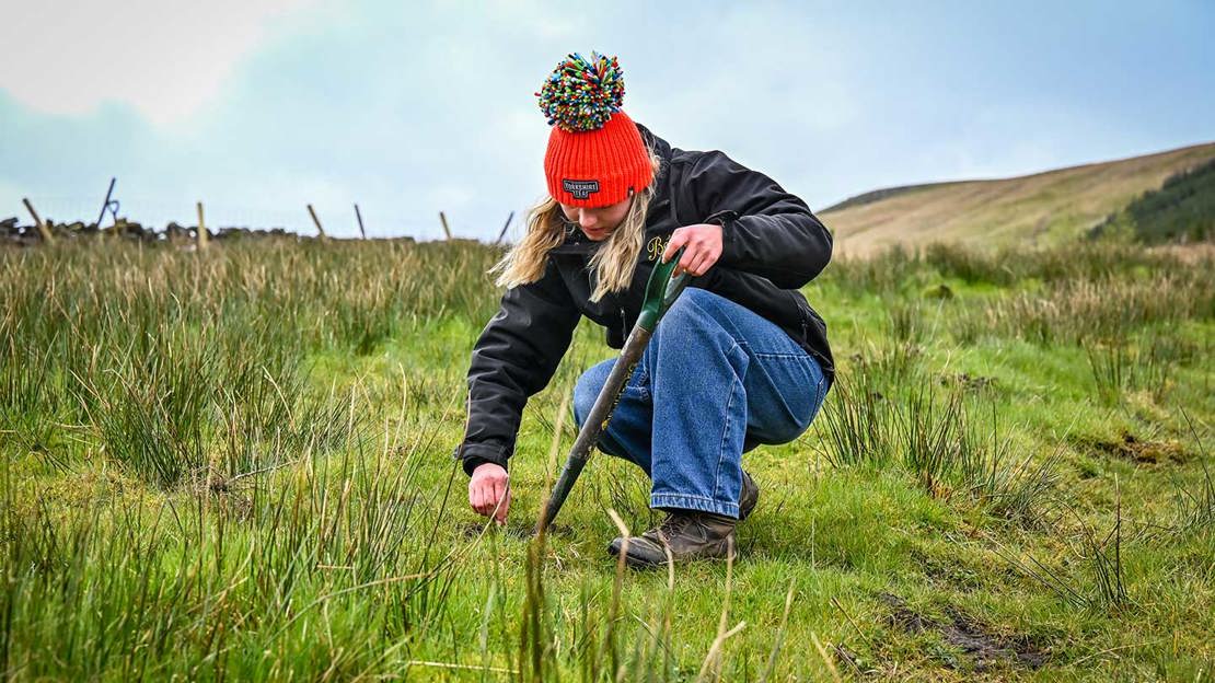 A young woman dressed in waterproofs and a red bobble hat digging into ground on the moors