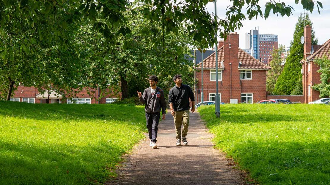 Two men walking under overhanging tree branches towards camera with houses in the background