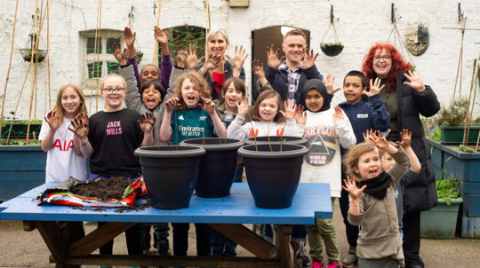 Author Lauren Child and school pupils standing behind plant pots and holding muddy hands up to the camera
