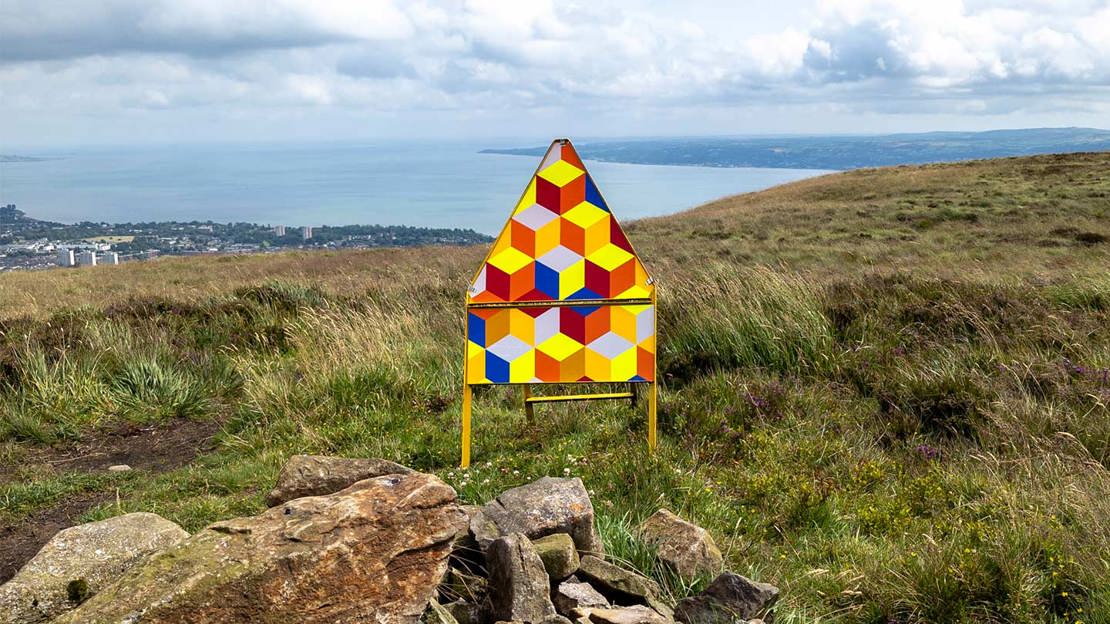 Triangular sign with colourful pattern in grass overlooking ocean.