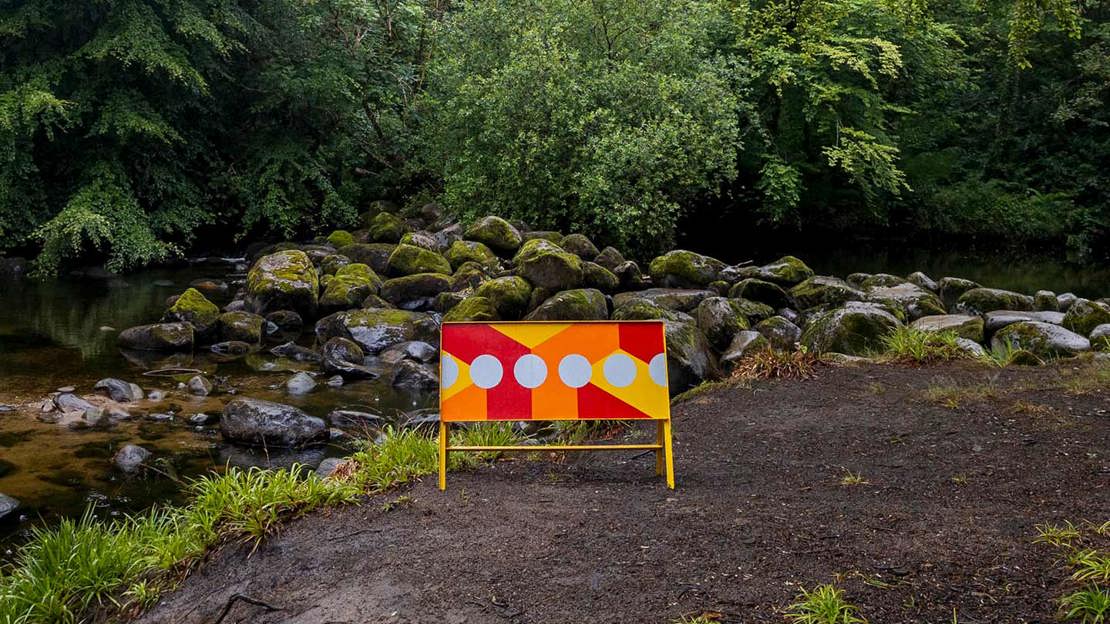 Rectangular sign with colourful pattern on woodland path in front of stream.