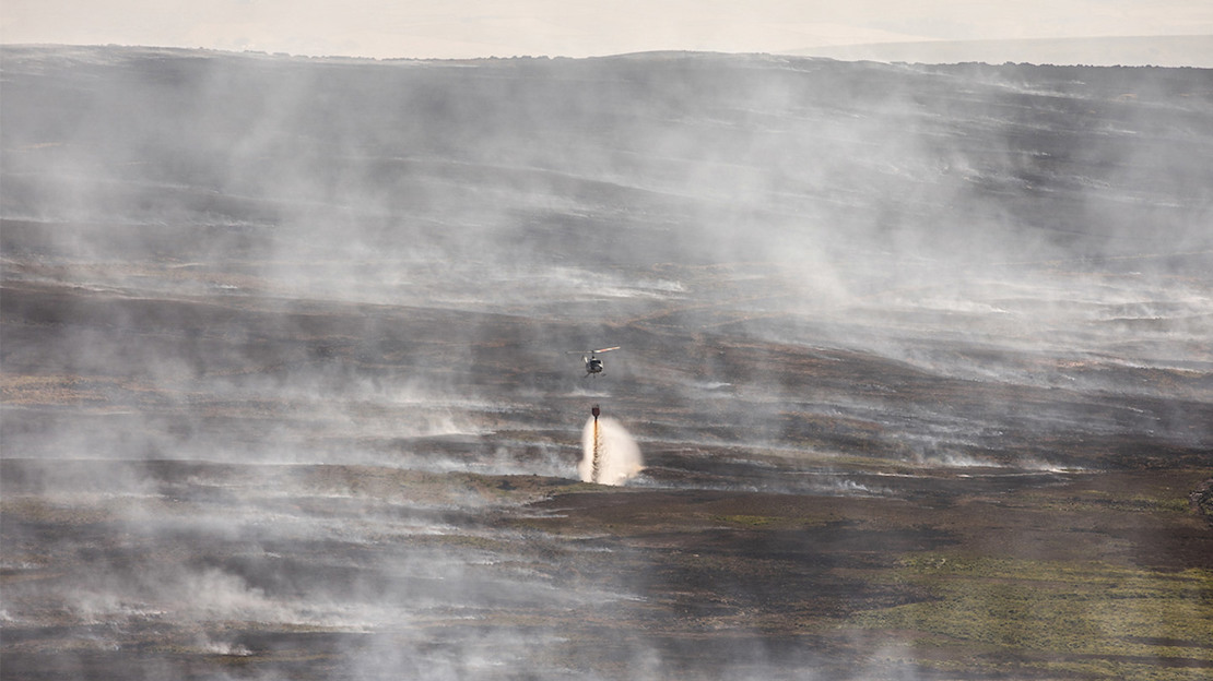 Firefighting helicopter dropping water over burnt landscape surrounded by smoke.
