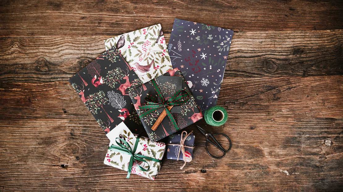 Three different sheets of nature-themed wrapping paper, two wrapped gifts and a pair of scissors on a wooden surface