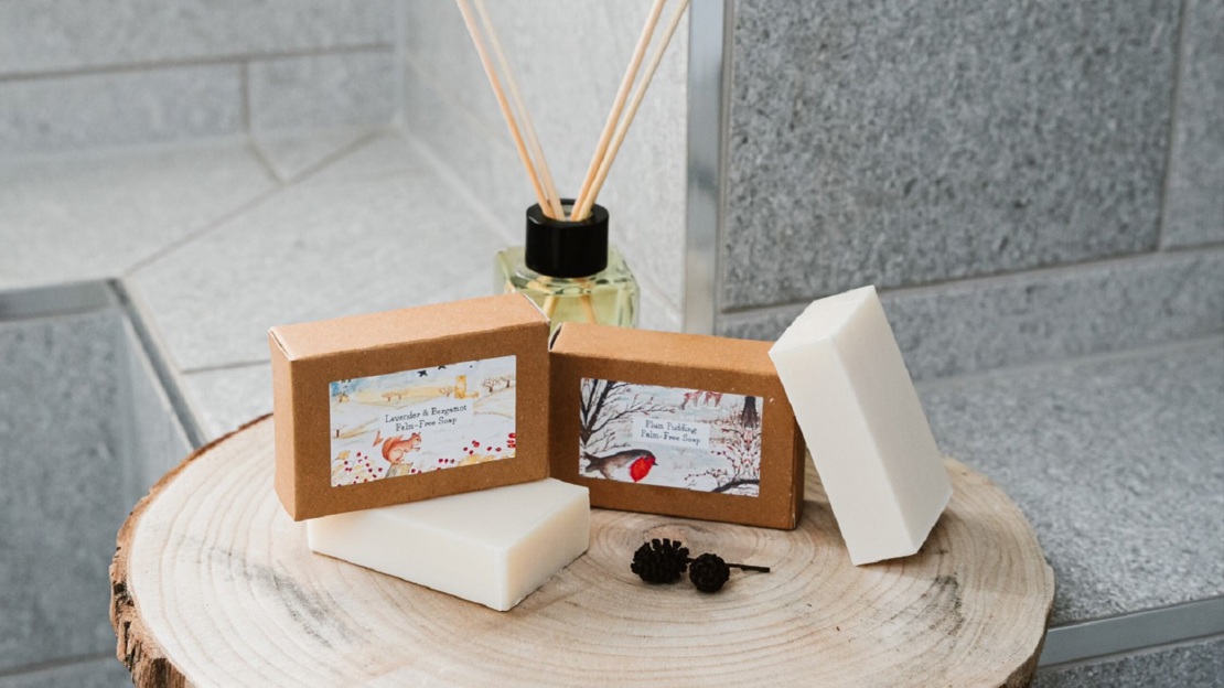 Two soaps arranged with their boxes and a reed diffuser in a grey tiled bathroom